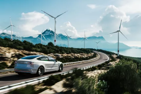 Ein silbernes E-Auto fährt bei blauem Himmel und Sonne eine Landstraße entlang. Rechts und links sind Büsche und Bäume.